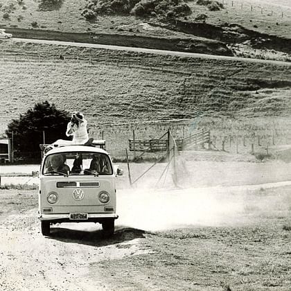 a kombi being driven on a dirt road with a photographer riding on the roof