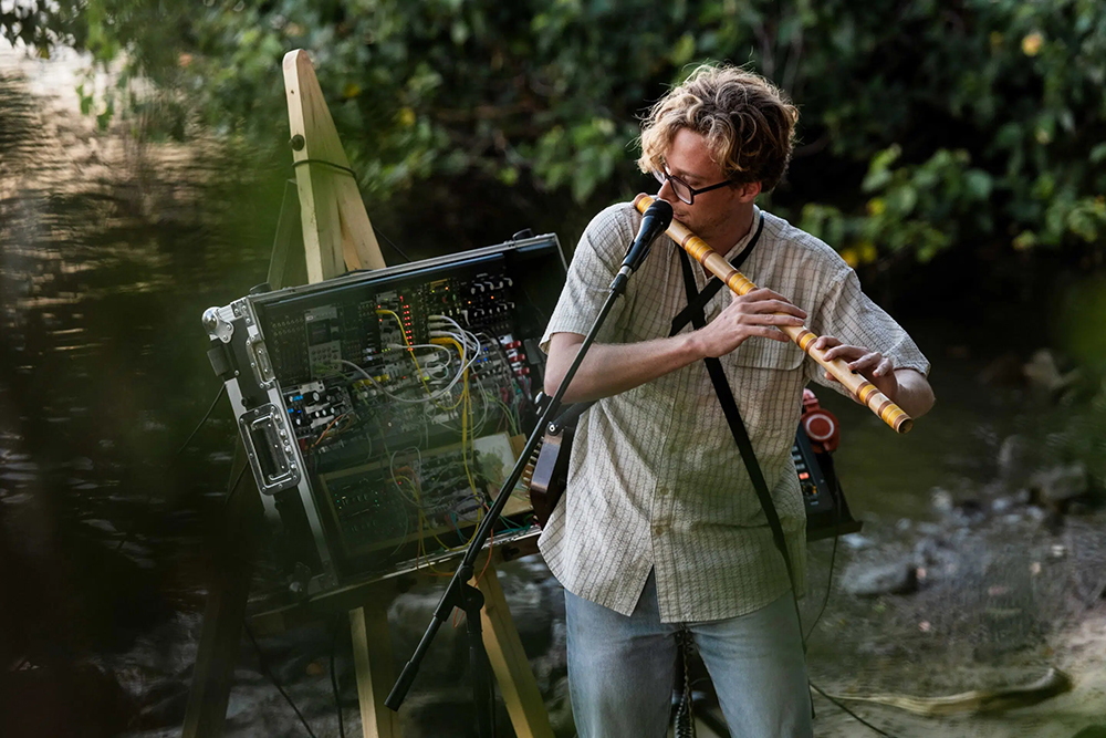 A young man playing a type of flute next to a river. There is a large electronic music making device as well. 