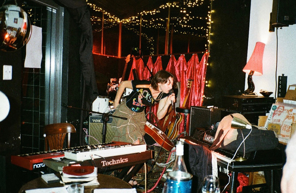 A woman singing into a microphone whilst holding a guitar and in front of a keyboard. There is a red curtain and fairy lights behind her in a cluttered room.