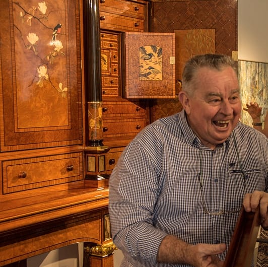 Geoff Hannah in front of the cabinet he crafted. Cabinet has inlays of timber and stone with many drawers and doors 