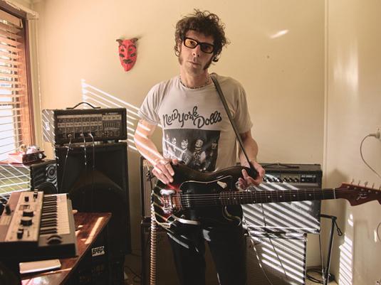 A man with sunglasses and a New Your Dolls t-shirt stands in a bedroom studio, holding a guitar and looking at the camera. You can see instruments behind him.