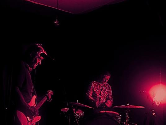 Two men playing in a darkened room with a crimson hue. One plays guitar, the other drums