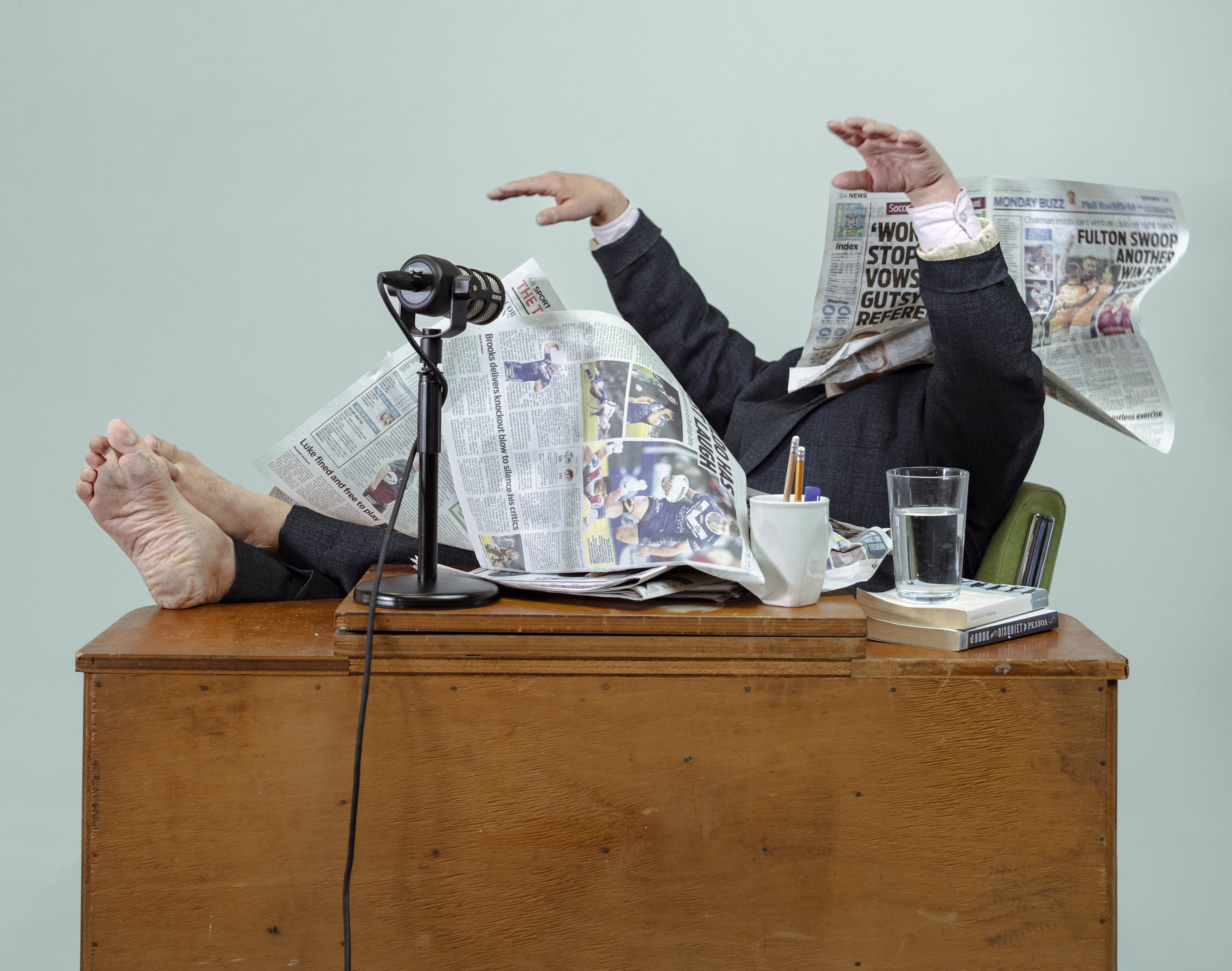 a person in a men's suit with feet on a timber desk, arms in the air and face and much of their body covered by newspaper pages.