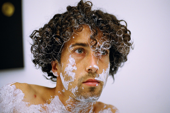 A portrait of a man with curly hair and white paint over his face and body