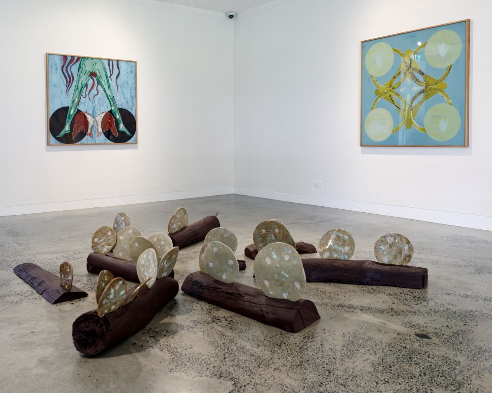 A exhibition install with 2 paintings on the wall and a wood and ceramic floor installation. There are repeating circular motifs, hues of blue, yellow, red and brown. 