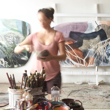artist in studio mixing paint with artworks behind her and jars of brushed in the foreground