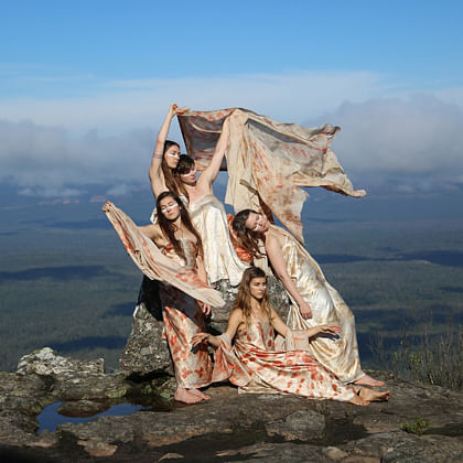 five women dancing with shawls on a high rock shelf with forest and lake landscape behind them
