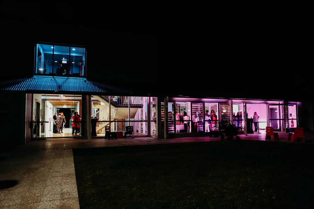 A view of a two storey building lit up at night with purple, blue and yellow nights. Taken from outside. 