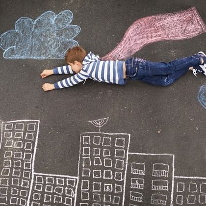 child lying on ground with drawings of building below them and a cloak drawn on ground so it appears he is wearing it and flying over the buildings and under the cloud