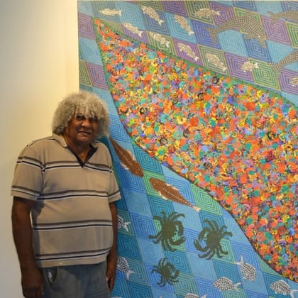 Digby Moran standing next to one of his paintings that includes a bright colourful island in the middle with fish and crabs swimming around it on a background of intricately patterned squares 
