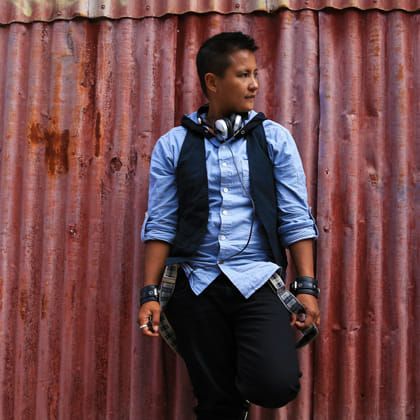 person in blue shirt with dark vest and trousers standing in front of corrugated iron shed