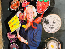 Photo of Peggy PopAr. She has blonde hair, a bright smile with red lips , a red scarf and hat. She is holding a sign and behind her are cutout images including a strawberry, a roller skate, a fried egg in a pan, a white cut out with smiling lips, one eye and a cylinder over the other eye and