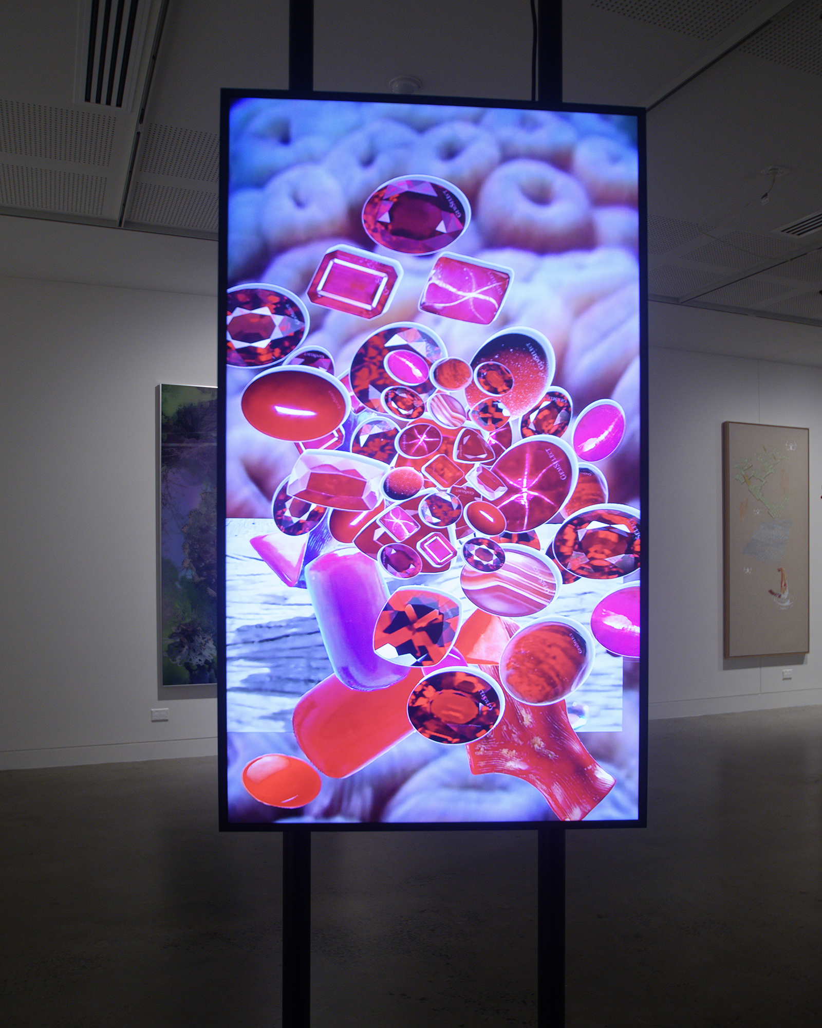 An exhibition documentation photo of a vertical screen showing a video with bright pink and purple jewel like objects floating