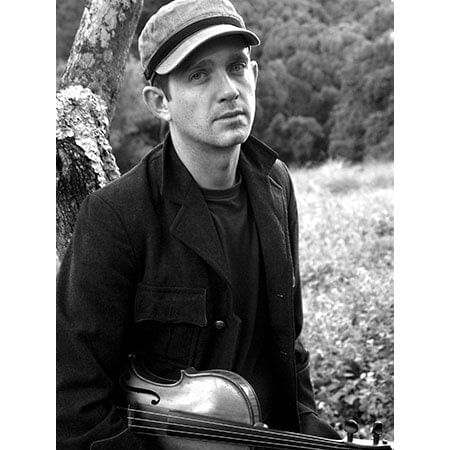 Cye Wood holding with a violin under his arm in front of a tree trunk, a  field and forest in background
