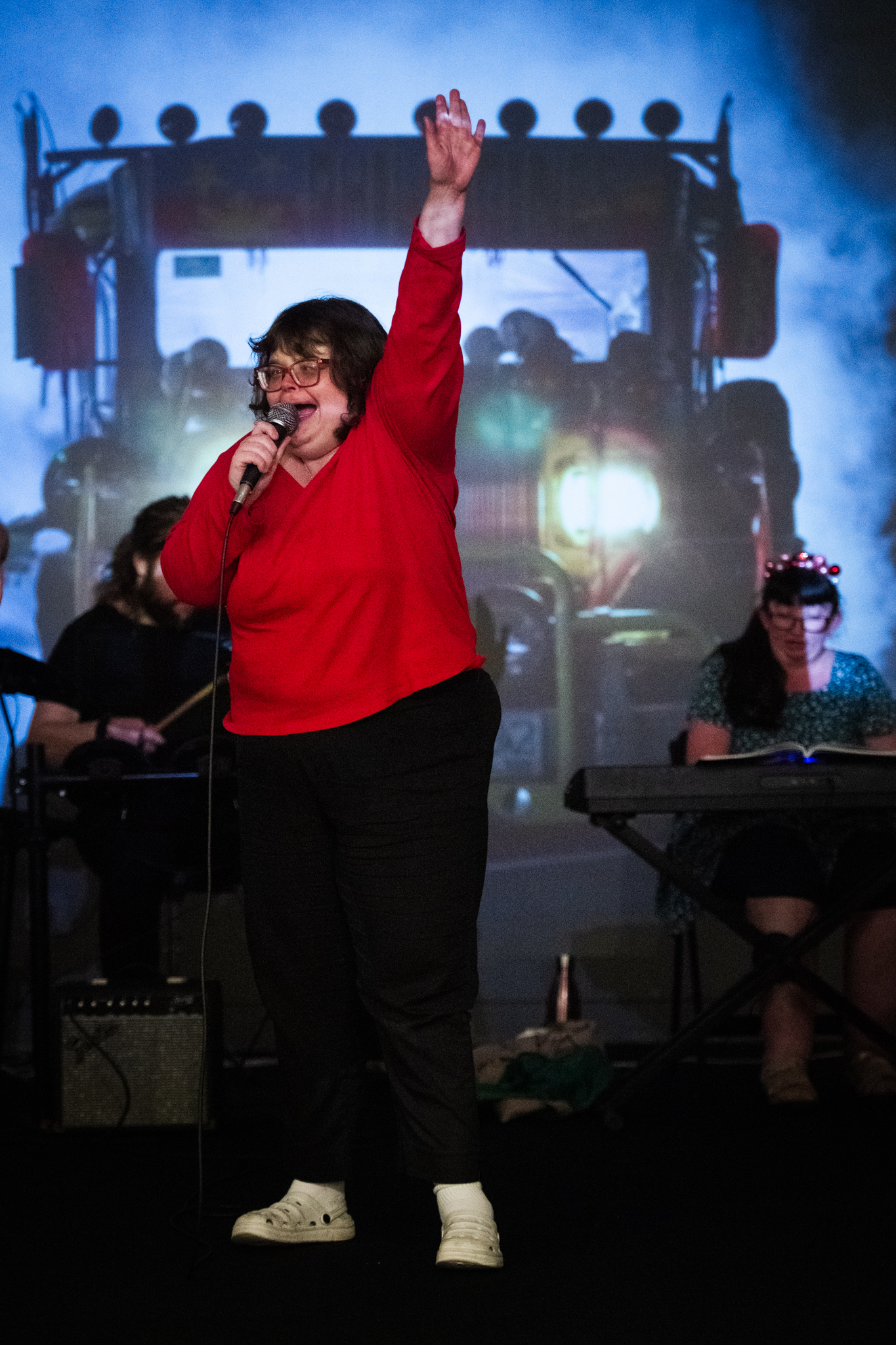 A woman with mixed abilities singing into a microphone with an arm raised in joy, in front of a projection