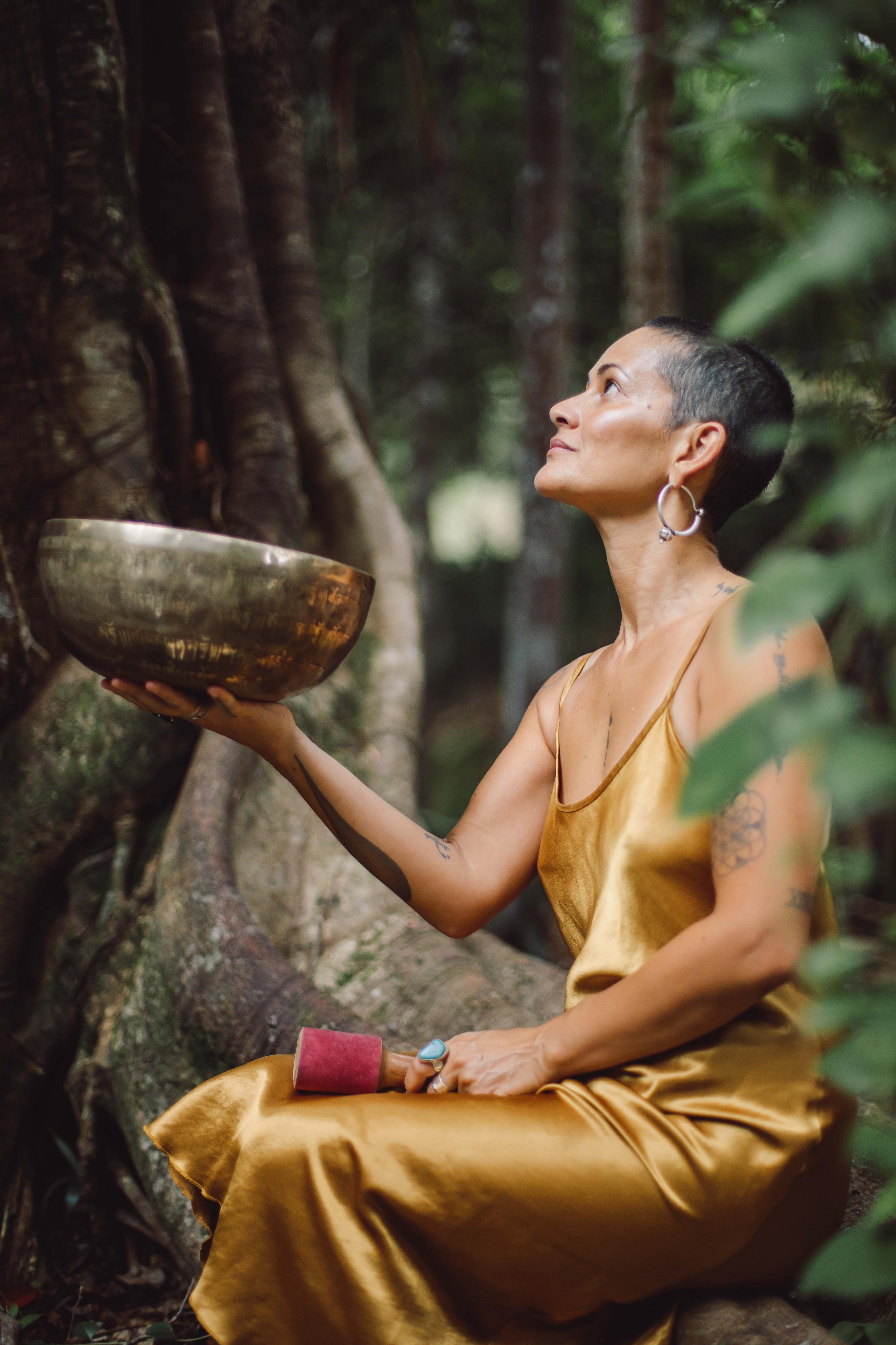 A woman of colour in a gold dress holding a singing bowl and looking up at the rainforest surrounding her