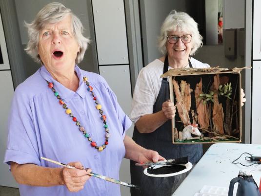Two smiling senior women holding their assemblage artworks