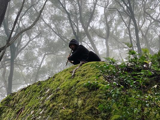 A person wearing all black crouched on a mossy rock, playing flute, with a misty, forest background behind them