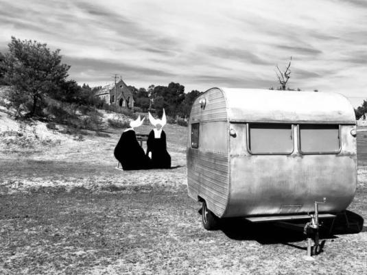 A black and white photo of a vintage caravan with two nuns in the background