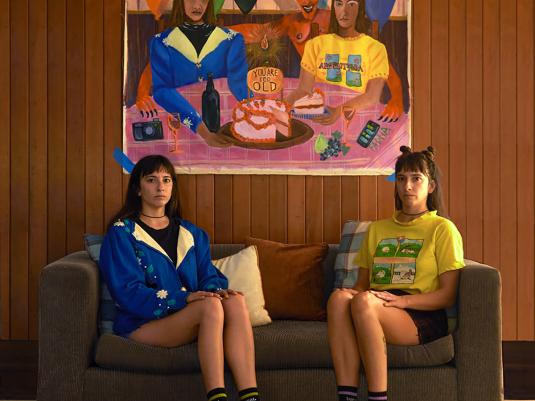 The artist in double, seated on two sides of the couch, with a self-portrait painting hanging above, echoing the double self below.