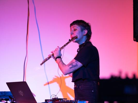 A non-binary person playing the flute, wearing black, with pink stage lighting