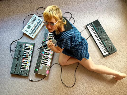 A woman with a blonde mullet haircut sitting on the floor surrounded by keyboards