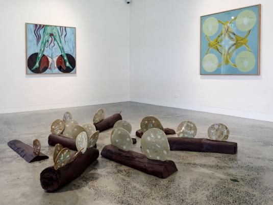 A exhibition install with 2 paintings on the wall and a wood and ceramic floor installation. There are repeating circular motifs, hues of blue, yellow, red and brown.