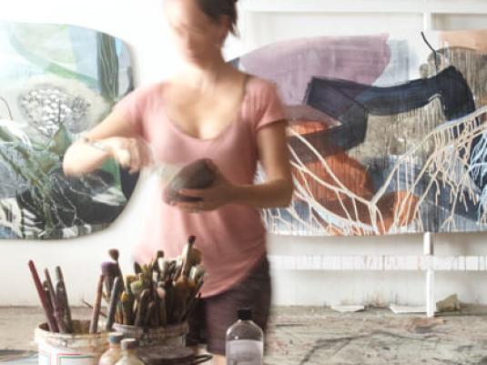 artist in studio mixing paint with artworks behind her and jars of brushed in the foreground