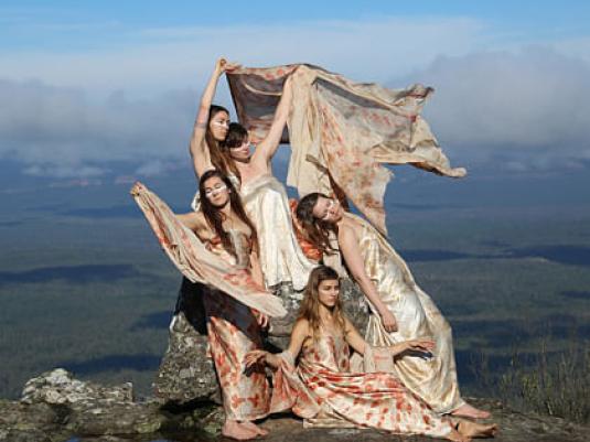 five women dancing with shawls on a high rock shelf with forest and lake landscape behind them