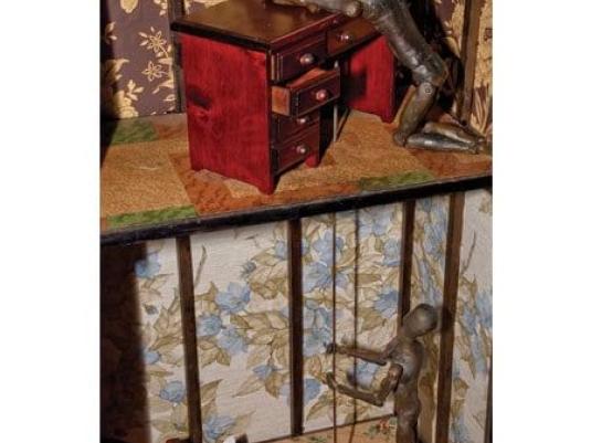 assemblage construction with one human form on top level and another below pulling on a string that goes up to the form on the top level