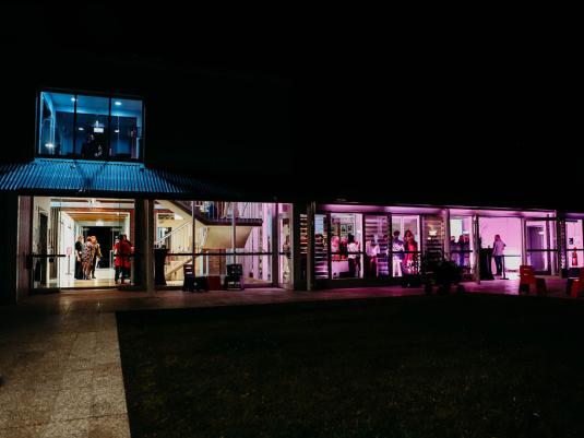 A view of a two storey building lit up at night with purple, blue and yellow nights. Taken from outside.