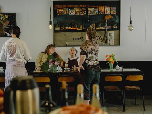 4 people in a Cafe, two standing, with their backs facing the camera, and two seated, laughing and animated. The group are of mixed gender, size and age. A large artwork is behind them and the table has many delcious dishes of food and flowers.