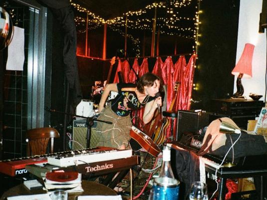 A woman singing into a microphone whilst holding a guitar and in front of a keyboard. There is a red curtain and fairy lights behind her in a cluttered room.