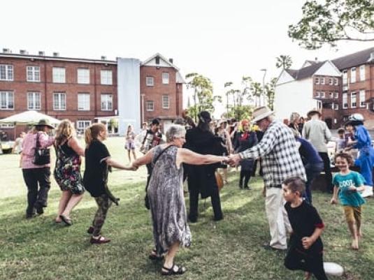 group of people in the gallery Quad. Some children running around, spome people holding hands and dancing in a circle with other  people are gathered in background e