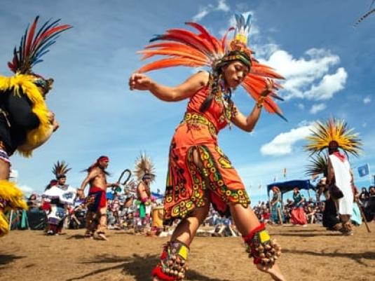 woman in Sioux tribal dress with headdress dancing in foreground with other people dancing in background in tribal dress.