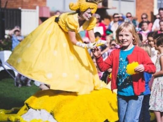 a crowd of children behind a woman dressed in yellow frock with wide skirt with a yellow headdress giving a child somethin yelloww