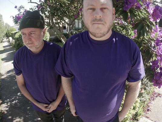 Two men in blue shirts and black jeans in front of a flowering vine.